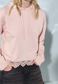 Růžová mikina s kráceným designem, která má krajkový lem na spodním okraji. Dlouhé rukávy a kapuce na šňůrku dotvářejí ležérní vzhled.