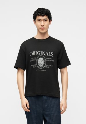 Ung mann iført en svart T-skjorte med "ORIGINALS" og et fjellmotiv med tekst, kombinert med mørkeblå bukser, stående mot en hvit bakgrunn.