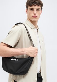 Jeune homme portant une chemise beige à manches courtes, tenant un sac bandoulière noir BOSS avec un logo blanc, regardant avec confiance vers la caméra sur un fond uni.