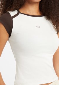 Tielko v off-white farbe s krátkymi hnedými rukávmi a výstrihom. Na prednej strane sa nachádza logo "GUESS JEANS" v tmavom písmene.