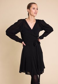 Svart omslagskjole laget av lett materiale, med lange pufferm, dyp V-utsnitt og midjebånd for justerbar passform.