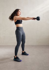 Melnā sporta krūšturis un augstās jostasvietas legingi ar teksturētu tumšu rakstu. Modelis tur kettlebellu, tērpusies melnos sporta apavos ar purpura akcentiem.