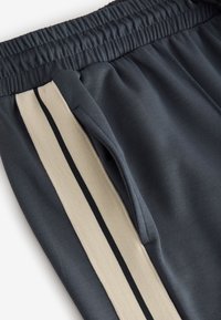 Tummanharmaat urheiluhousut, joissa on kuminauhavyötärö, sivutasku ja beige sivuraidallinen mustilla yksityiskohdilla. Sileä pinta.