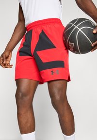 Man in rode en zwarte Under Armour basketbalshorts, houdt een zwart-witte Spalding basketbal vast en draagt witte sokken.