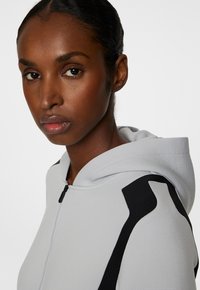 Ljusgrå zip-hoodie med strukturerad tyg, svarta detaljer på axlarna och en justerbar huva.