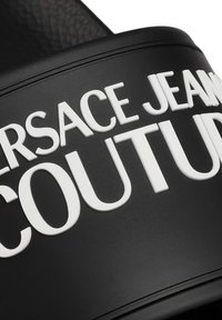 Sandale noire à brides avec texte en relief de couleur blanche indiquant "VERSACE JEANS COUTURE." Le texte est en gras, avec une finition mate texturée.
