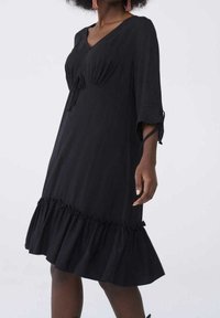 Svart knälång klänning i mjukt tyg, med v-ringning, volangnederdel och 3/4-ärmar med knyt vid ärmsluten.