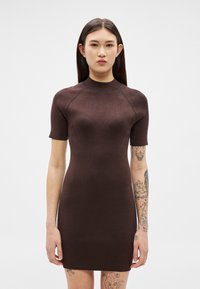 Γυναίκα με ίσια σκούρα μαλλιά που φοράει εφαρμοστό, ριμπ, καφέ φόρεμα με κοντά μανίκια, εμφανίζοντας τα�τουάζ στα μπράτσα και τα πόδια της, πάνω σε λευκό φόντο.