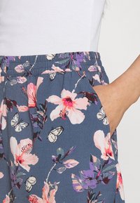 Tela azul con estampado floral de flores rosas y moradas, con cintura elástica y bolsillos laterales. Bolsillo parcialmente visible.