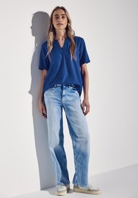 Blusa blu a maniche corte con scollo a V, abbinata a jeans in denim azzurro. Il modello indossa sneakers beige. Lo sfondo è bianco uniforme.