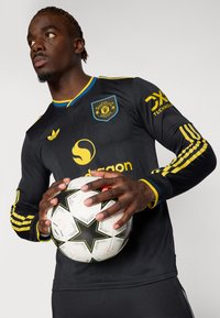 Black long-sleeve soccer jersey with yellow accents, featuring a logo and details. Player holds a black and white star-patterned soccer ball.