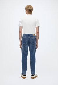Joven con el pelo rubio corto que lleva una camiseta blanca lisa, vaqueros azules y zapatillas blancas, de espaldas.