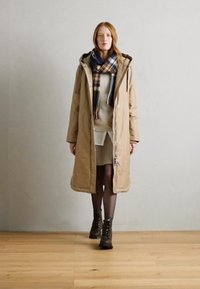 Beige dlouhý zimní kabát s kapucí, nošený přes šedý svetr a krémové šaty. Tmavě kostkovaná šála a hnědé šněrovací boty dotvářejí outfit.