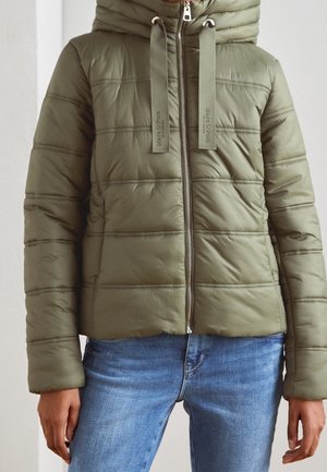 Olivengrøn quiltet dunjakke med hætte, lynlås og mærkeindtastede snore, båret sammen med blå denimjeans.