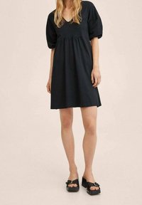 Vestido corto negro con mangas abullonadas y escote en V, con cintura fruncida y corte holgado. Combinado con sandalias negras de plataforma.