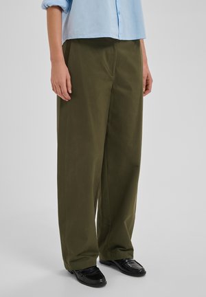 Persoon draagt losse olijfgroene broek, zwarte loafers en een lichtblauw shirt met lange mouwen, staand op een effen lichtgrijze achtergrond.