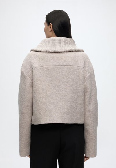 Femme aux cheveux raides et foncés porte un pull court beige clair avec un large col côtelé et des manches longues, associé à un pantalon noir, vue de dos.