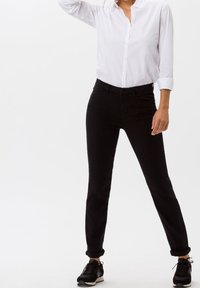 Jeans negros ajustados de talle medio, con cinco bolsillos y una textura suave, combinados con una camisa blanca de botones y zapatillas negras.