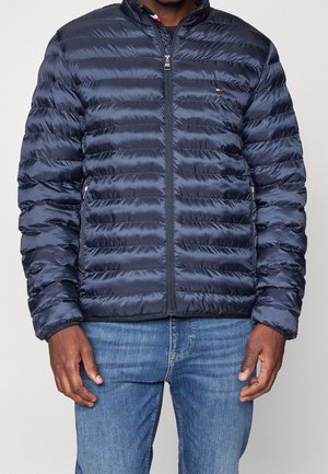 Homme portant une doudoune matelassée bleu foncé avec poches zippées et fermeture éclair à l'avant, associée à un jean bleu, visible du cou jusqu'aux hanches.