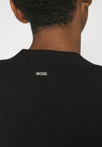 Gros plan sur le cou et le haut du dos d'une personne portant un vêtement noir avec un petit logo argenté "BOSS" à l'encolure.