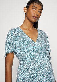 Vestido envolvente teal com decote em V, mangas onduladas e um padrão floral branco. O tecido é macio com um acabamento ligeiramente texturizado.