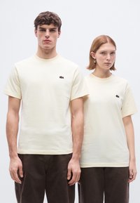 Ανοιχτό κίτρινα βαμβακερά μπλουζάκια με κοντά μανίκια και στρογγυλή λαιμόκοψη. Και τα δύο έχουν ένα μικρό σκοτεινό λογότυπο στο στήθος. Απλός σχεδιασμός, άνετη εφαρμογή.