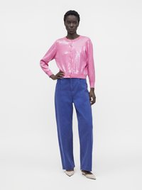 Femme debout, main sur la hanche, portant un cardigan rose brillant boutonné, un pantalon bleu ample et des chaussures pointues de couleur claire sur fond blanc.
