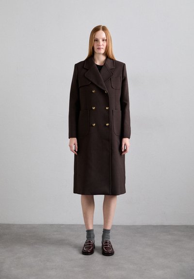 Manteau marron à double boutonnage avec de grands revers, boutons dorés et deux poches avant. Porté sur une robe noire, associé à des chaussettes grises et des mocassins.