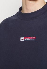 T-shirt in cotone blu navy con colletto rotondo a coste, caratterizzato da un patch rettangolare del logo Tommy Jeans in rosso, bianco e blu sul petto.