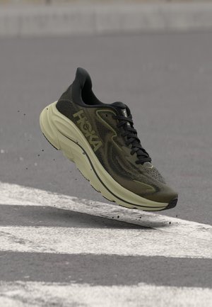 Oliivinvihreä ja musta Hoka-juoksukenkä roikkuu harmaan asfaltin yläpuolella, jonka alla on valkoisia tienviivoja ja pieniä mustia roskia.