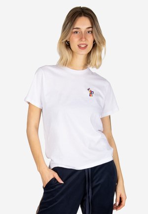 Frau mit schulterlangen blonden Haaren trägt ein weißes T-Shirt mit kleinem buntem Motiv und dunkle Hose, steht mit einer Hand in der Tasche.