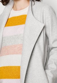 Grijze wollen jas met revers over een zachte gebreide trui, met horizontale strepen in geel, roze, grijs en crème kleuren. Geëffekteerde stof.