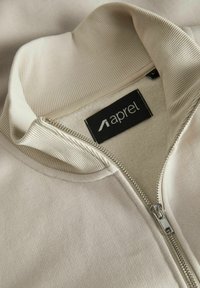 Light beige zip-up jacket with ribbed collar, soft lining, and branded tag. Features a silver zipper and smooth exterior texture.