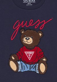 Mornarska majica od organskog pamuka s teksturiranim, izvezenim smeškom smešnog smešnog smeša koji nosi crveni duksericu s trokutastim logotipom i plave traperice.