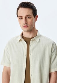 Joven con cabello corto y oscuro que lleva una camisa de botones de manga corta beige sobre una camiseta de punto marrón, frente a un fondo claro y sencillo.