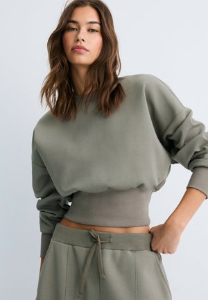 Junge Frau mit braunen Haaren, die ein lockeres, olivgrünes cropped Sweatshirt und passende Hosen mit Kordelzug trägt, posiert mit einer Hand auf der Hüfte.