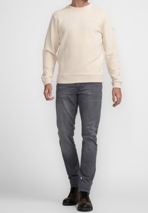 Mann trägt beigen Rundhalspullover, graue Jeans und schwarze Schnürstiefel und steht vor einem schlichten Hintergrund.