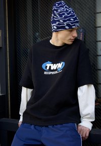 Mladý muž nosiaci modro-bielu vzorovanú čiapku, čierne tričko s logom TWN cez biele tričko s dlhým rukávom a modré nohavice, sediaci opretý o sklenené dvere.