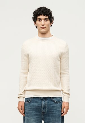 Jeune homme aux cheveux bouclés et foncés, portant un pull crème à torsades et un jean bleu, debout devant un fond clair uni.