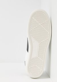 Sula av sko med ett vitt gummimaterial, hexagonalt strukturerat mönster, släta svarta läderdetaljer och en halvcirkelformad kantdesign.