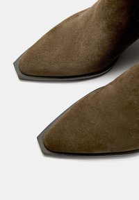 Bottines en daim marron avec orteils pointus et semelles en caoutchouc noires. Présentent une texture lisse et des lignes géométriques nettes.