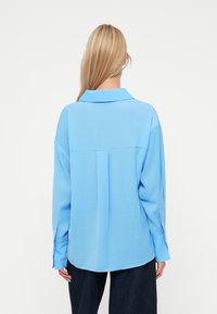 Ανοιχτό μπλε πουκάμισο με κουμπιά, φτιαγμένο από λείο ύφασμα, διαθέτοντας γιακά, μακριά μανίκια και ελαφρώς καμπυλωτό εντύπωμα, με λεπτομέρειες πτυχής στην πλάτη.