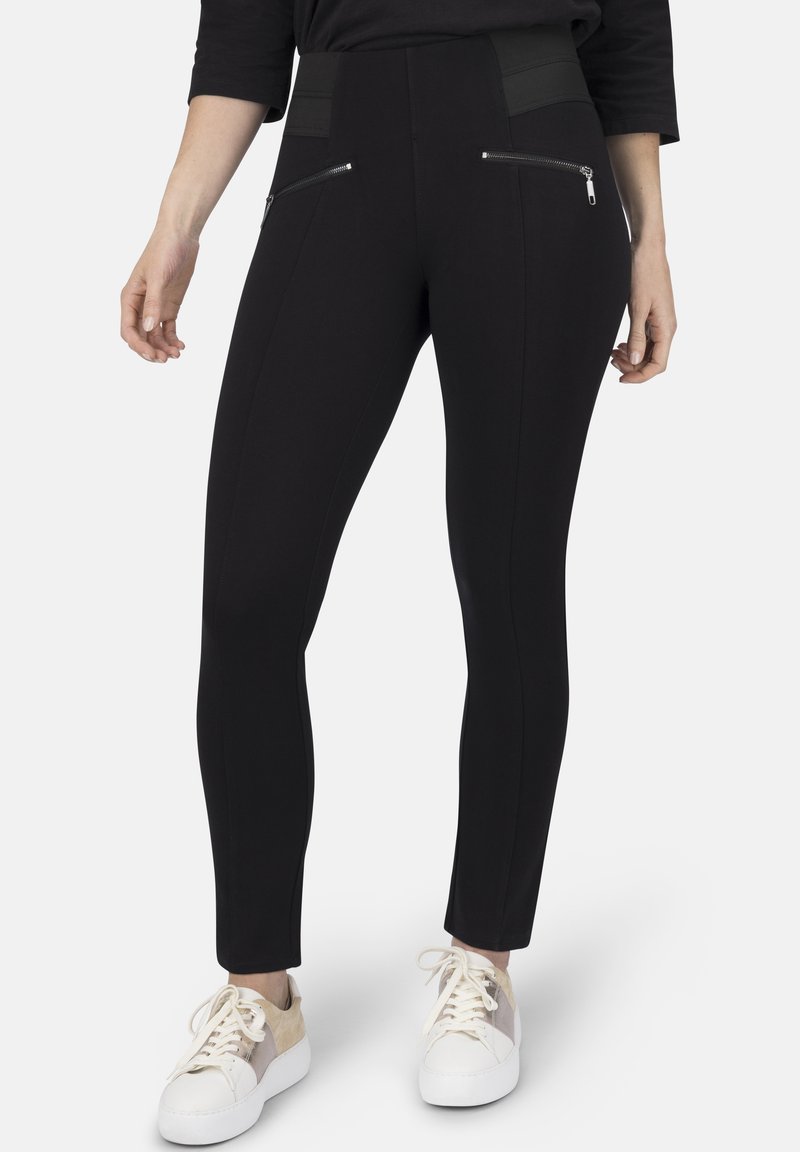 Schwarze Stretch-Leggins mit hohem Bund, zwei Reißverschluss-Vordertaschen und kontrastierenden Texturpaneelen an den Seiten. Kombiniert mit weißen Sneakers.