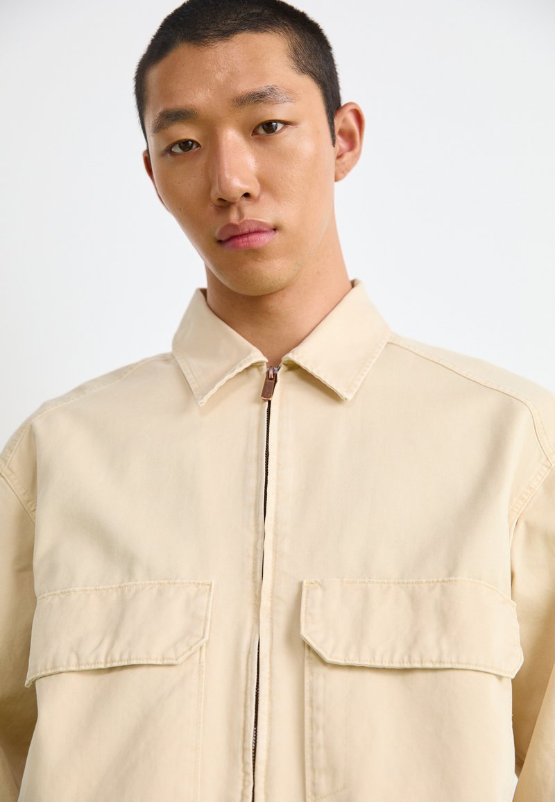 Veste en toile beige avec fermeture éclair sur le devant, deux poches poitrine et un col pointu. Présente des coutures détaillées et une surface texturée.