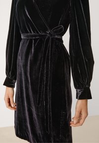 Svart sammet svepande klänning med långa puffärmar, knytmidja och en slät yta som framhäver tygets lyster.