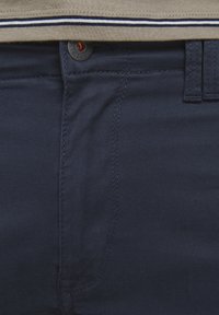 Pantalon bleu marine avec bouton visible et couture de fermeture éclair, associé à une chemise beige ornée d'une bande blanche et noire près de l'ourlet.
