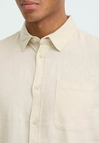 Chemise beige boutonnée en tissu texturé. Elle présente un col pointu, une poche plaquée et des boutons contrastants sur le devant.