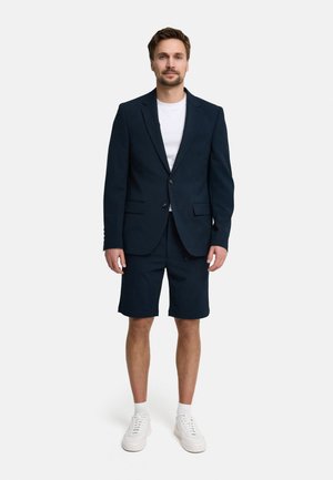 Hombre de pie vestido con un blazer azul marino, pantalones cortos a juego en azul marino, camiseta blanca, calcetines blancos y zapatillas blancas sobre un fondo claro liso.