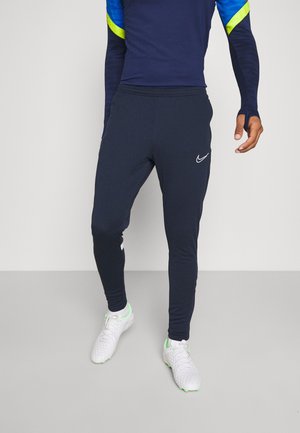 Homme portant un pantalon de sport Nike bleu marine, des chaussures de sport blanches et un haut bleu à manches longues avec des accents jaune néon, debout sur un sol gris.