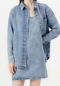 Light blue denim shirt over a matching denim dress. The shirt features a button-up front, pocket, and slightly oversized fit.
